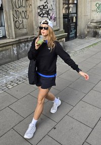 Svart sweatshirt med blå detaljer, svarta shorts, vita sneakers och håller en grön dryck. Solglasögon och en vit keps fulländar outfiten.