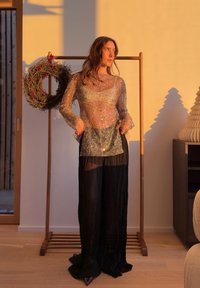 Femme portant un haut transparent scintillant et un pantalon large noir, debout à l'intérieur près d'un cadre en bois avec une couronne de fête, baignée de lumière chaude.