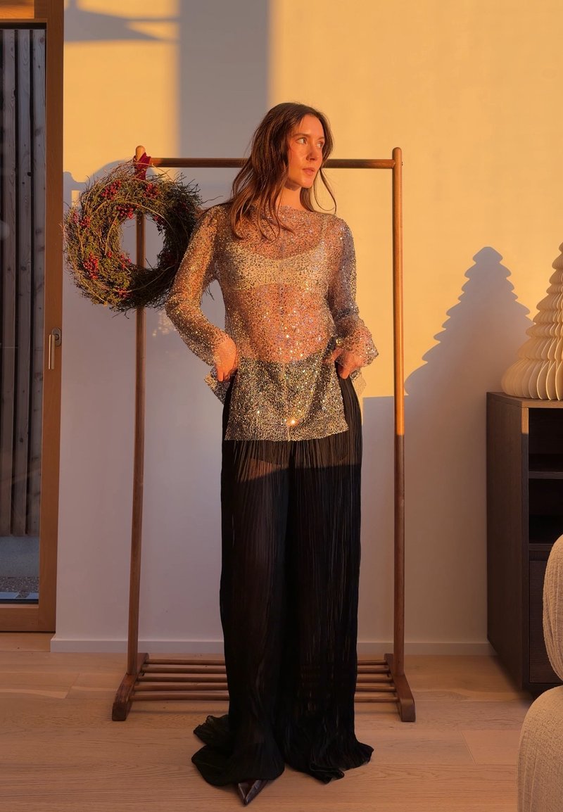 Femme portant un haut transparent scintillant et un pantalon large noir, debout à l'intérieur près d'un cadre en bois avec une couronne de fête, baignée de lumière chaude.