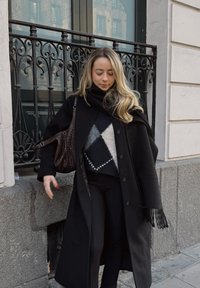 Manteau noir à gros boutons, porté par-dessus un pull à motifs en noir, gris et blanc. Pantalon foncé et sac marron texturé avec une sangle.