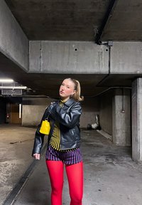 Νεαρή γυναίκα που ρυθμίζει μαύρο δερμάτινο μπουφάν, φοράει κίτρινο ριγέ πουκάμισο, μωβ ριγέ σορτς, κόκκινες καλτσοδέτες, στέκεται σε υπόγειο πάρκινγκ με τσιμεντένιο δάπεδο.
