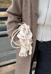 Biely látkový vak s volánovými okrajmi a vyšívaným hnedým vzorom. Drží ho ruka, doplnený o hnedý huňatý kabát a svetlý sveter.