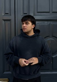 Νεαρός άνδρας με κοντά σκούρα μαλλιά, φορώντας μια μαύρη φούτερ με κουκούλα, στέκεται μπροστά από μια σκοτεινή πόρτα με πάνελ, κοιτάζοντας προς το πλάι.