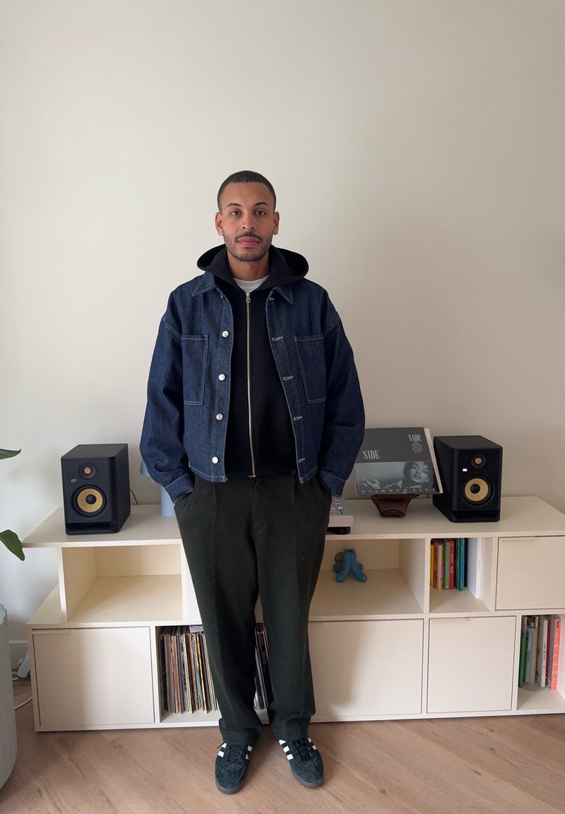 Homme portant une veste sombre, un sweat à capuche et un pantalon, debout les mains dans les poches devant une étagère contenant des enceintes, des disques et des livres.