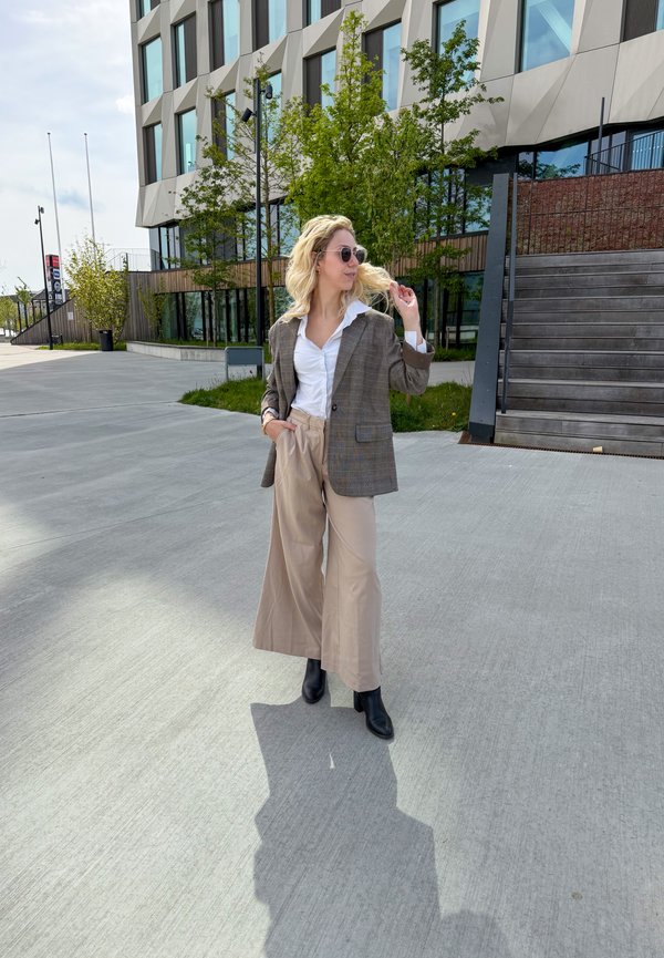 Frau mit Sonnenbrille, braun kariertem Blazer, weissem Hemd und beiger weiter Hose steht auf Beton in der Nähe eines modernen Gebäudes mit Bäumen und Treppen.