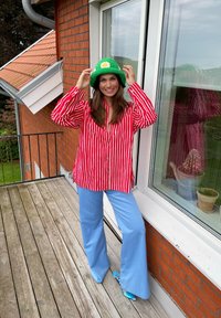 Camicia oversize a righe rosse e bianche, pantaloni svasati azzurri chiari, cappello a secchiello verde all'uncinetto con un fiore bianco e ciabatte turchesi.