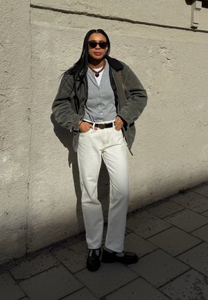 Modelo con una chaqueta verde oliva sobre un cárdigan gris, jeans blancos y zapatos negros de punta redonda. Los accesorios incluyen collares en capas y gafas de sol.