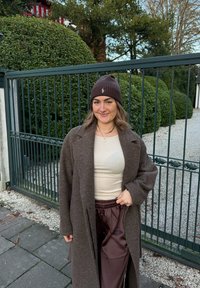 Brun ullfrakk, beige tettsittende topp og brune satengbukser. Antrekket kompletteres med en mørk brun lue med logodetalj.