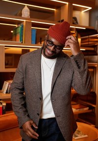 Veste en tweed marron portée sur un t-shirt blanc, jean bleu et bonnet en tricot orange côtelé. Lunettes noires et collier pendentif en argent.