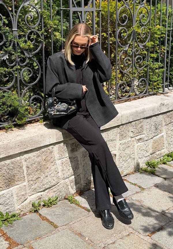Žena oblečená v tmavom oversized saku a nohaviciach sedí na kamennej stene pri dekoratívnom železnom plote a upravuje si slnečné okuliare v slnečný deň.