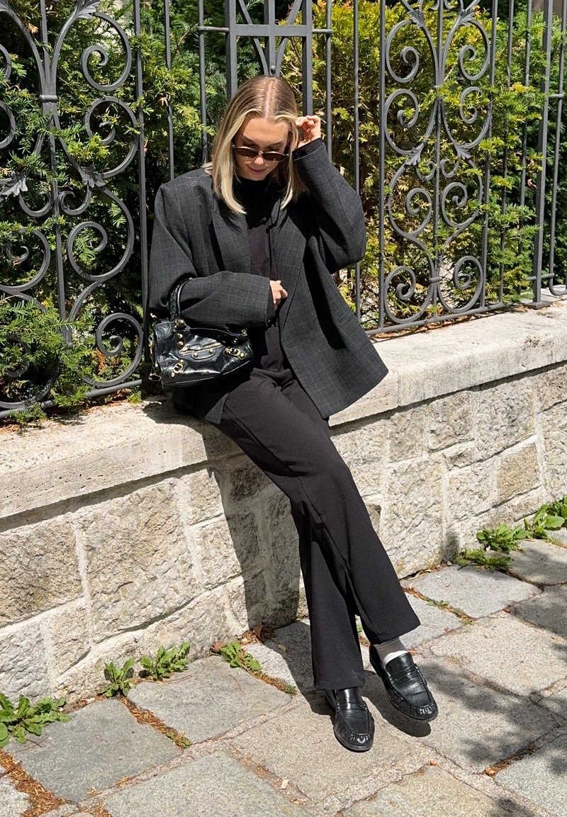 Femme portant un blazer sombre oversize et un pantalon, assise sur un mur de pierre près d'une clôture en fer décorative, ajustant ses lunettes de soleil par une journée ensoleillée.
