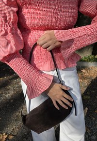 Schwarze Wildleder-Handtasche, die in der Hand gehalten wird, kombiniert mit einem rot-weiß karierten, gerafften Oberteil. Es hat ausgestellt Ärmel und weiße Hosen.