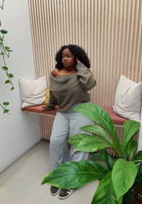 Maglione oversize verde oliva con design off-shoulder, pantaloni a gamba larga grigio chiaro e sneakers verdi. Lo sfondo presenta cuscini morbidi e pannellature in legno.