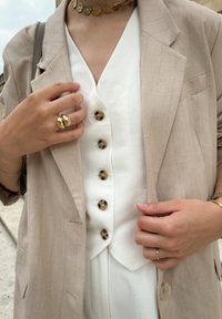 Beige gestreepte blazer over een witte blouse met bruine knopen, aangevuld met gouden sieraden, waaronder een choker en ring.
