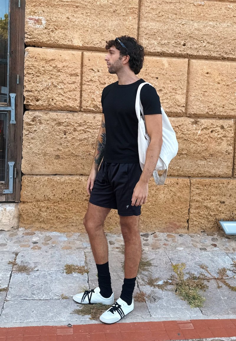 Maglietta nera a maniche corte, pantaloni neri, scarpe bianche con lacci neri e una borsa tote bianca, in piedi contro un muro di pietra texturizzato.