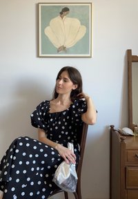 Rochie neagră cu buline albe, cu mâneci bufante, ținând o geantă metalică argintie. În fundal, un scaun din lemn și artă înrămată. Lumină naturală.