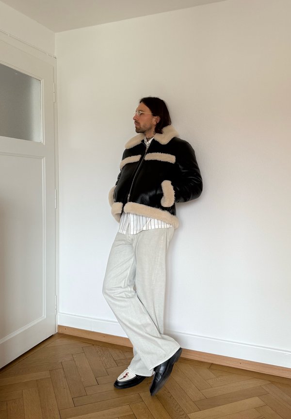 Mand iført sort shearlingjakke, stribet skjorte, hvide bukser og loafers, der læner sig op ad en hvid væg i et rum med trægulv.