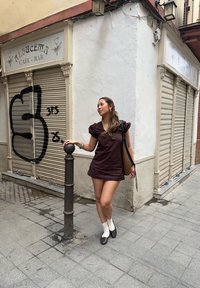 Robe courte marron à manches bouffantes, associée à des chaussettes blanches et des ballerines marron. Un sac tissé pend de l'épaule, sur un fond urbain texturé.