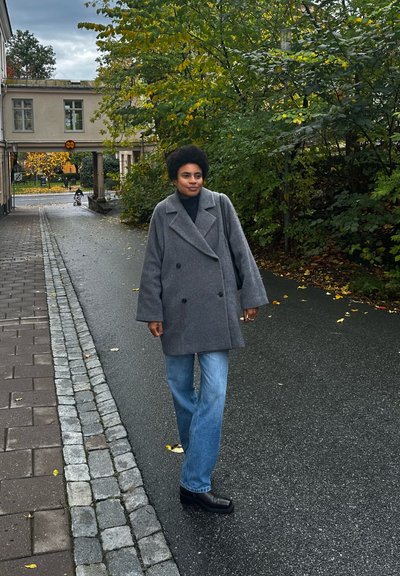 Abrigo de lana gris de doble botonadura, vaqueros anchos azules y botas negras. La escena incluye hojas en el suelo y un camino de piedra.