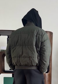 Personne portant une veste matelassée verte et une capuche noire, vue de dos, debout devant une étagère en bois contenant des livres et un carnet.