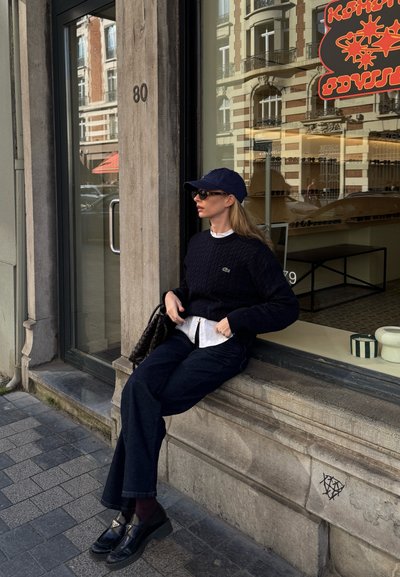 Suéter de punto trenzado azul marino, camisa blanca de cuello, jeans oscuros, mocasines negros con suela gruesa y una gorra azul marino a juego. Sentado en un borde de piedra.