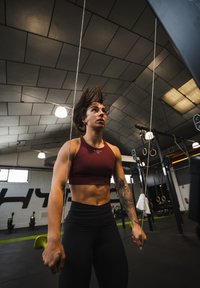 Atlet iført en mørkerød crop top og sorte leggings, som laver øvelser med træningselastikker i et fitnesscenter med højt til loftet og udstyr.