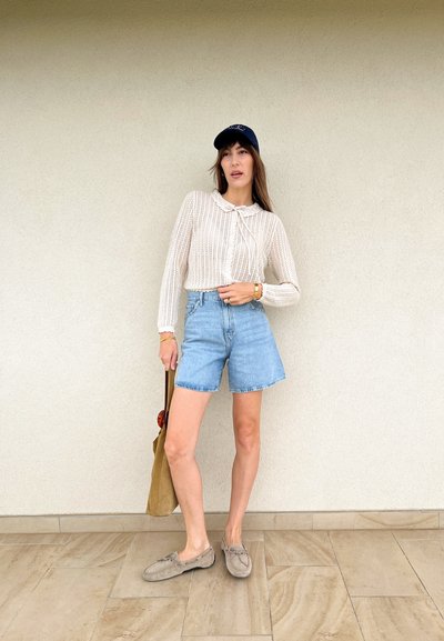 Jersey de punto crema con cuello, pantalones cortos de mezclilla azul claro, zapatos mocasín beige y una bolsa de tote beige, todo ello frente a una pared lisa.