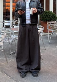 Svart skinnjakke over en hvit skjorte, kombinert med løstsittende, mørkegrå bukser. Personen holder en smarttelefon og en kaffekopp. Svarte sko.