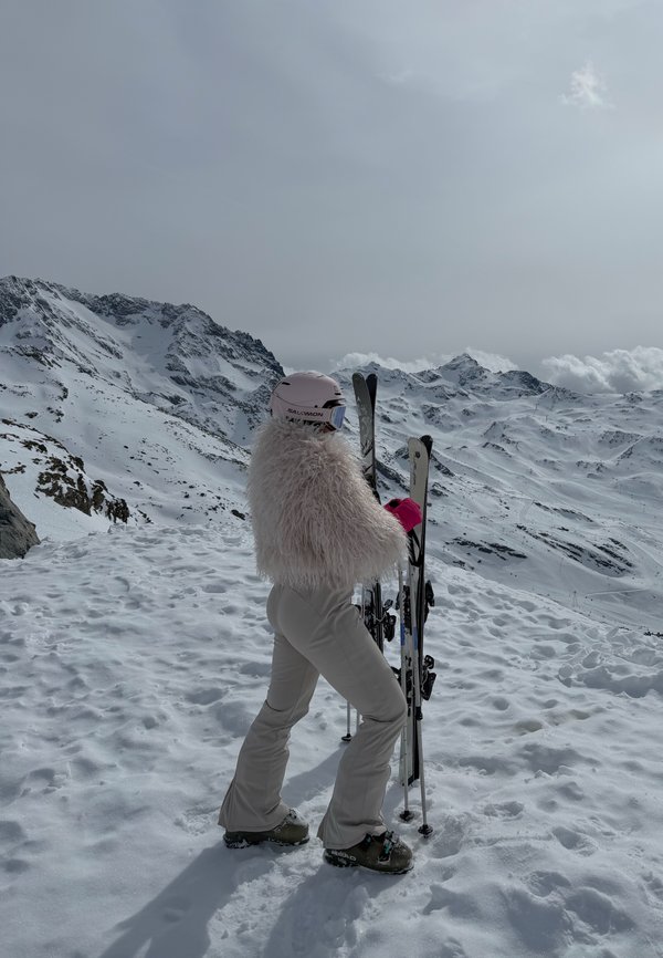 Persoon die een wit skipak en helm draagt en ski's vasthoudt, staat op een besneeuwd berglandschap onder een bewolkte lucht.