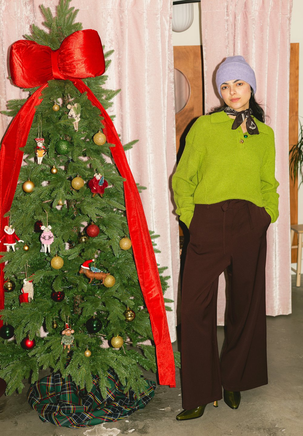 Femme portant un pull vert, un bonnet violet et un pantalon marron, debout à côté d'un sapin de Noël décoré avec un grand nœud rouge et diverses décorations.