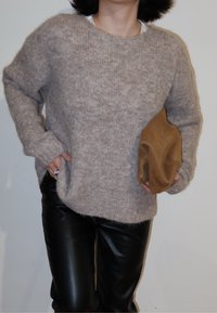 Personne portant un pull beige en tissu pelucheux et un pantalon en cuir noir, tenant un chapeau en daim marron plié dans une main, contre un mur uni.