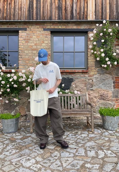 Hombre con gorra azul y camiseta blanca está junto a un banco, sosteniendo una bolsa de tela Marc O’Polo Denim, con flores y una casa de ladrillo al fondo.
