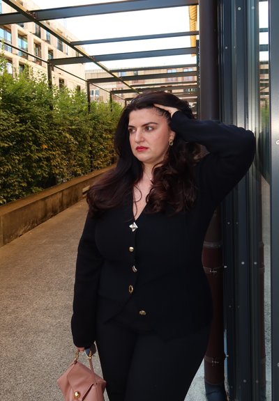 Cárdigan negro con botones dorados, combinado con una blusa y pantalones negros. Sosteniendo un bolso rosa claro con detalles dorados. Follaje verde de fondo.