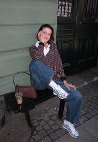 Felpa a righe bordeaux sopra una camicia bianca con colletto, jeans blu, scarpe grigie, con una borsa marrone, seduto su una panchina nera.