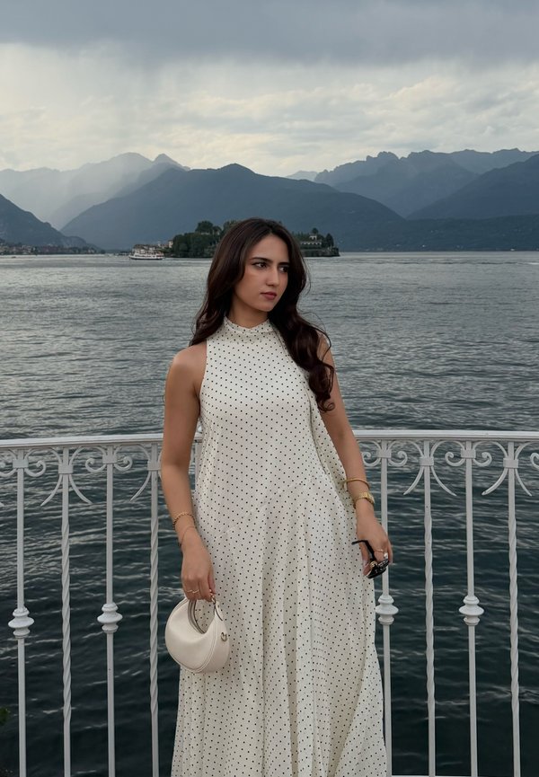 Mujer con vestido blanco de lunares que sostiene un pequeño bolso blanco y gafas de sol, parada junto a una barandilla blanca con un lago y montañas de fondo.