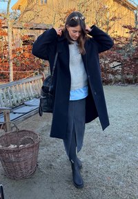 Manteau en laine noir porté sur un pull gris clair et une chemise bleue, assorti à une jupe en denim et des bottines noires. Accessoires : lunettes de soleil.