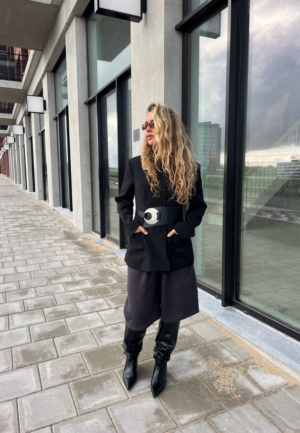 Blazer nero con tasche sopra un outfit scuro, stretto con una cintura larga in argento. Stivali slouch neri fino al ginocchio completano il look.