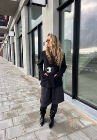 Blazer nero con tasche sopra un outfit scuro, stretto con una cintura larga in argento. Stivali slouch neri fino al ginocchio completano il look.