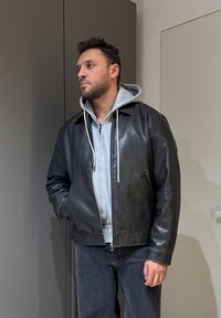 Veste en cuir noir avec un col rond, fermeture éclair et poches latérales, superposée sur un sweat à capuche gris. Portée avec un jean foncé.