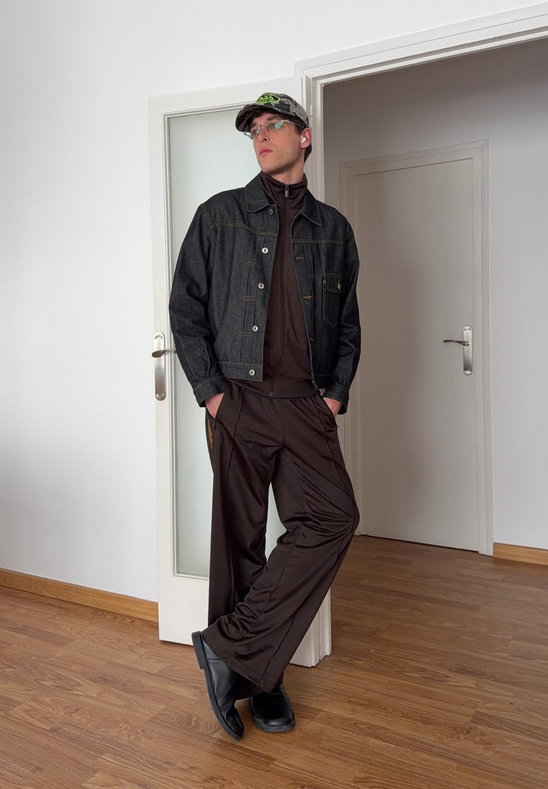 Jeune homme portant une veste en jean, un survêtement sombre, une casquette, des lunettes et des chaussures noires, appuyé contre un chambranle dans une pièce avec un sol en bois.