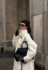 Valkoinen villakangastakki korkealla kauluksella ja mustilla napilla, yhdistetty mustaan laukkuun ja kapeisiin mustiin hansikkaisiin. Musta baretti ja aurinkolasit täydentävät asun.