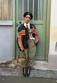 Musta ja oranssi nahkatakki, valkoinen paita ja pilkkuliina; vihreä baretti, cargo-housut, mustat nauhakengät ja seeprahäntä laukku. Vihreä oven tausta.