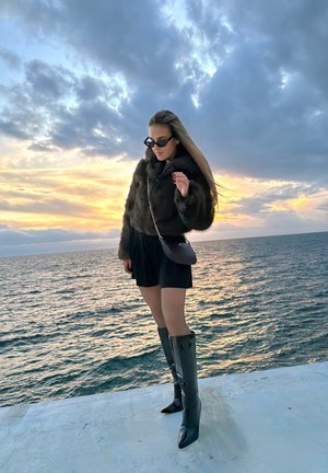 Mujer con gafas de sol, chaqueta de piel, falda negra y botas hasta la rodilla, de pie en una plataforma de hormigón junto al mar al atardecer con cielo nublado.