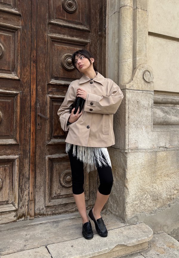 Femme en manteau beige et leggings noirs tenant une pochette noire, debout devant une grande porte en bois avec un mur en pierre décoré en arrière-plan.