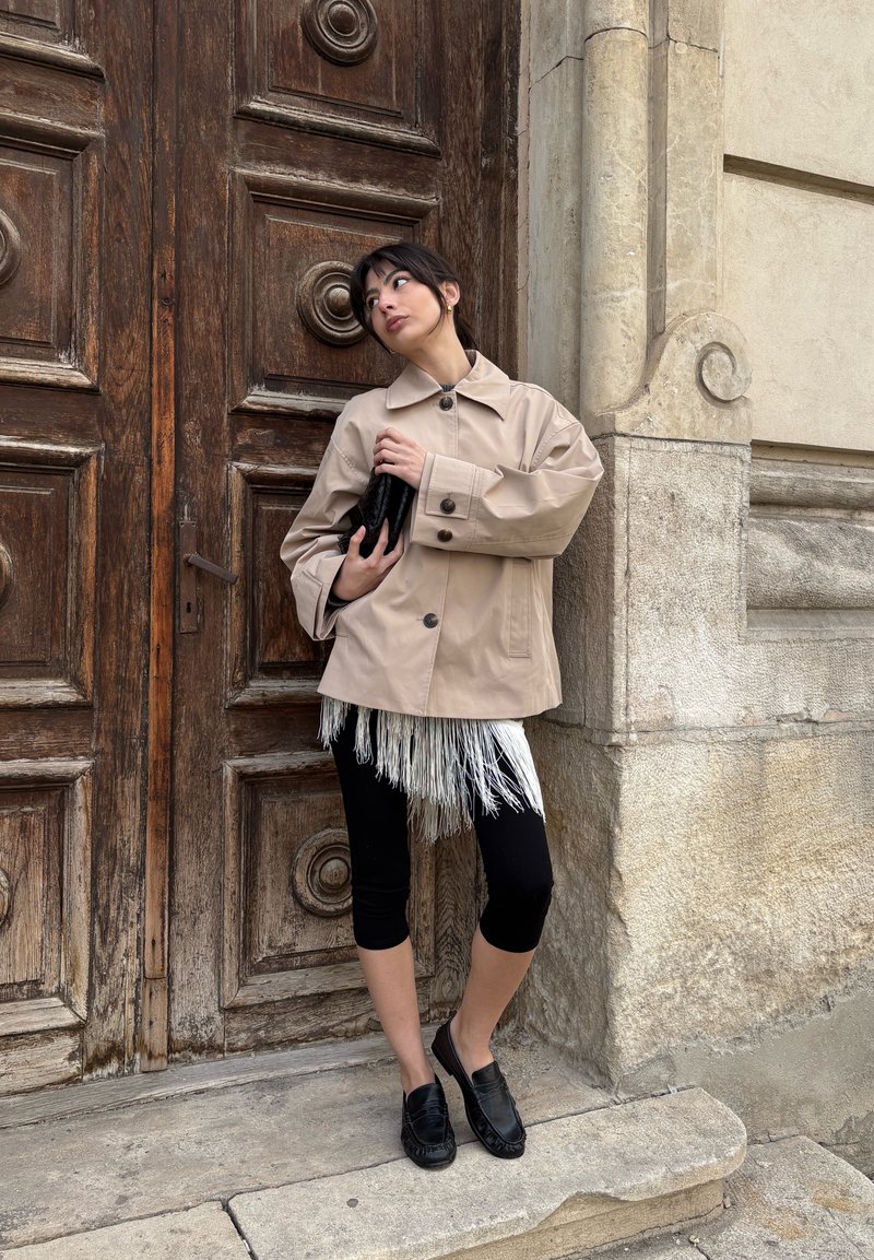 Femme en manteau beige et leggings noirs tenant une pochette noire, debout devant une grande porte en bois avec un mur de pierre orné en arrière-plan.
