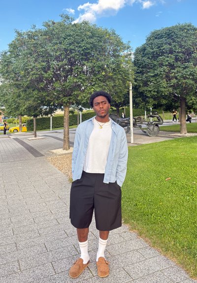 Una persona lleva una camisa de cuadros azul claro, una camiseta blanca, pantalones cortos negros y zapatos de tela marrones. Está de pie sobre un pavimento gris cerca de hierba verde.