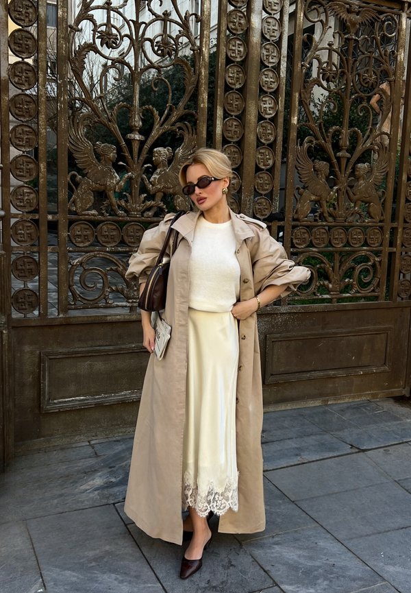 Nainen, jolla on beige trenssitakki, valkoinen neule, kerma satiinihame, jossa pitsireuna, tummat korkokengät, aurinkolasit ja ruskea käsilaukku koristellun portin edessä.