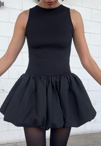 Donna che indossa un abito nero senza maniche con corpetto aderente e gonna ampia arricciata, in piedi contro un muro di mattoni bianchi.