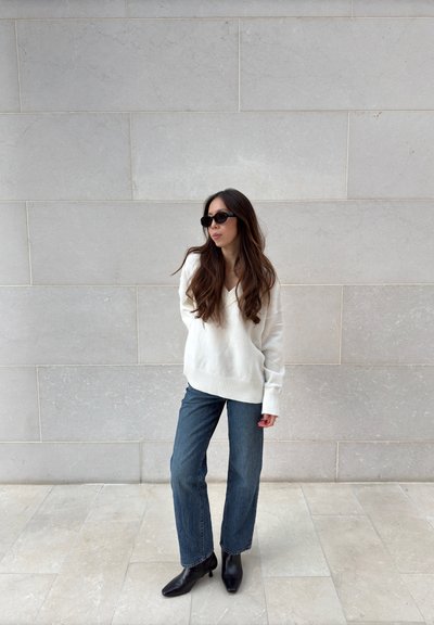 Suéter de punto blanco con escote en V, jeans de mezclilla azul de cintura alta y botines negros. El cabello largo y las gafas de sol completan el look.