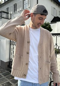 Béžový pletený kardigan s výstrihom do V, prednými gombíkmi a rebrovanými manžetami. Obsahuje dve predné vrecká a textúrovaný povrch. Nosený cez biele tričko.